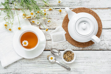 Cup of hot chamomile tea and teapot on light wooden background