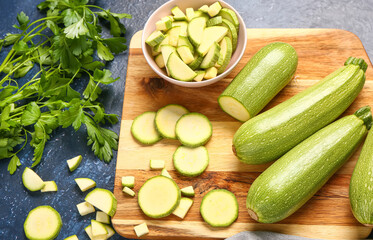 Board with fresh green zucchini and bowl of slices on blue background