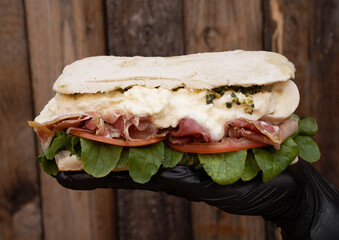 Chef wearing black gloves holding a gourmet sandwich made with ciabatta bread, prosciutto ham,...