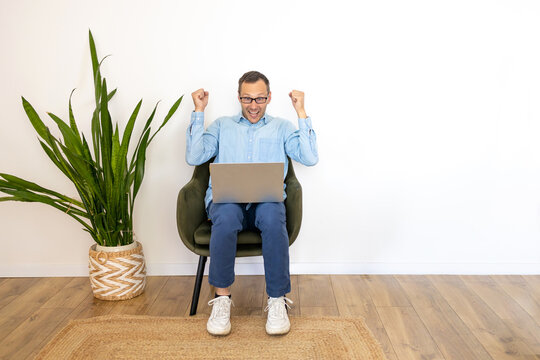 Emotional Young Man In Casual Sitting In Arm Chair With Laptop And Raising Hands Up, Happy Guy Got Dream Job Or Great News, White Empty Wall Background, Copy Space.