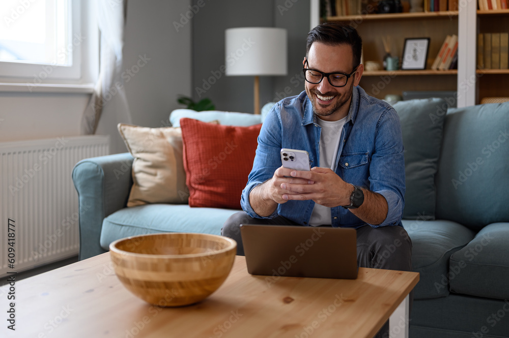 Wall mural Smiling young businessman with laptop on table text messaging over smart phone while sitting on sofa.Male entrepreneur telecommuting with wireless computer and cellphone while working from home office