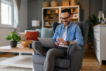 Confident young businessman smiling and analyzing strategies over laptop while sitting on armchair. Male freelancer making new business plans on wireless computer while working in home office