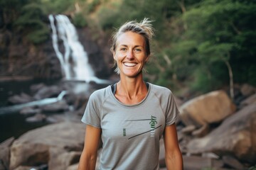 Medium shot portrait photography of a grinning woman in her 40s that is wearing a casual t-shirt against a scenic hiking trail with a waterfall in the distance background . Generative AI