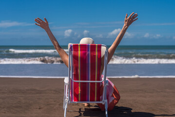 Woman raising her hands up while relaxing sitting on a deck chair at the beach.