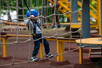 Summer. A small child climbs in a rope park on a rope bridge. A boy is having fun in an Adventure Park. A male baby on a climbing frame. Compliance with safety techniques.