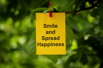 A yellow paper note with the words Smile and Spread Happiness on it attached to a tree branch with a clothes pin