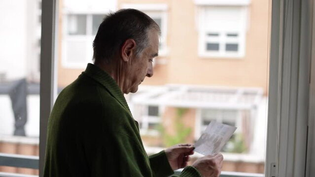 Senior man with Alzheimer's and depression, sideways, looking at a photograph he is holding, and looks out a window. Concept of old age, disease, loneliness, forgetfulness and memory.
