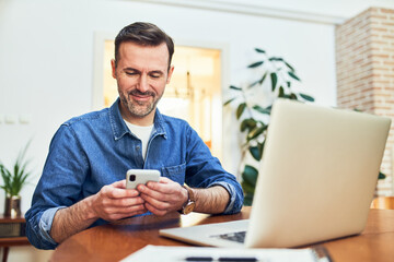 Smiling adult man using smart phone and laptop at home while working remotely