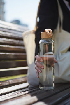 Young woman sitting on a bench in the morning and holding a glass water bottle in hand. Unrecognizable female person chilling outdoor in a sunny day with an eco bottle of fresh water