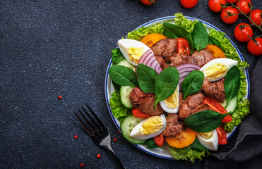 Perigord salad with roasted chicken liver, red tomatoes, cucumbers, boiled eggs, lettuce and spinach. Black kitchen table background, top view