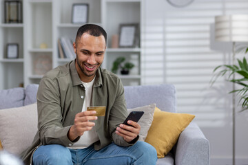Smiling young hispanic man sitting on sofa at home in hotel and using credit card and phone