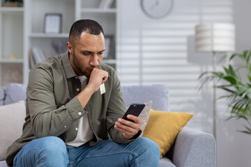 A young African American man received bad news, a message on his cell phone. He sits upset at home on the sofa, looks at the screen in shock