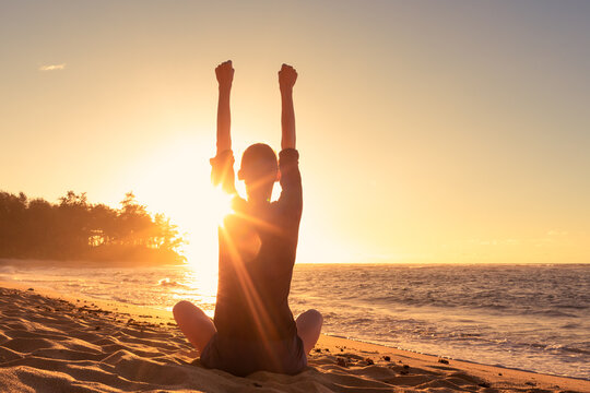 Young woman sitting on a beach feeling strong inspired energized facing the sunrise 