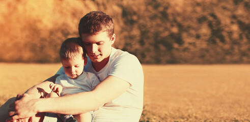 Portrait of happy young father and son child together sitting on grass in sunny evening park
