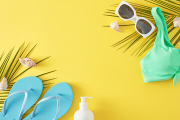 Concept of a tropical beach vacation. Top view flat lay of stylish teal swimsuit, flip-flop, sunglasses, sunscreen, palm leaves and seashells on light yellow background with area for text or ads