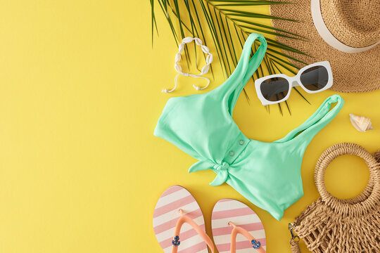 The idea of a tropical holiday. Top view flat lay of teal swimsuit, beach bag, straw hat, flip flops, sunglasses, shell bracelet, palm leaf, seashell on yellow background with space for promo or text