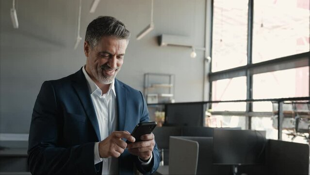 Happy middle aged business man ceo wearing blue suit standing in office using cell phone. Older businessman professional executive holding mobile satisfied with enterprise solution management service.