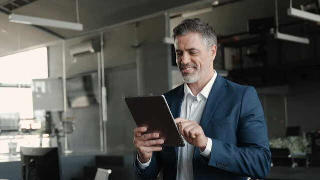 Smiling mature professional business man, happy middle aged successful company ceo, confident executive investor wearing blue suit standing in office holding tablet computer corporate technology.