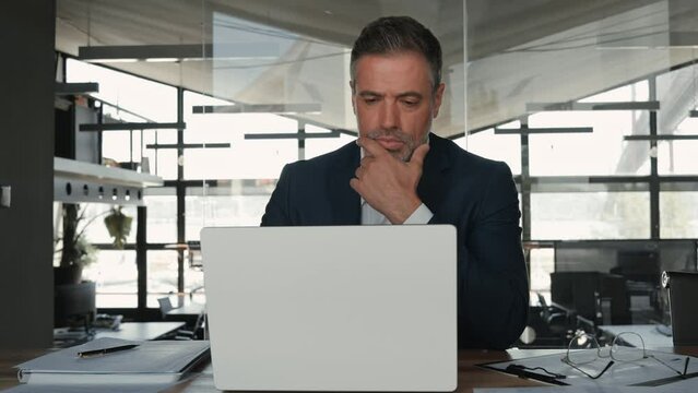 Busy mid aged business man ceo executive investor wearing suit looking at laptop computer analyzing financial data, managing corporate risks, thinking over project strategy solution working in office.