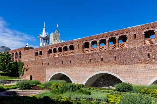 Red stone and brick bridge leading to Akhaltsikhe (Rabati) Castle, with cloisters and old moat overgrown with plants, flowers and grass, castle watchtower in the background.