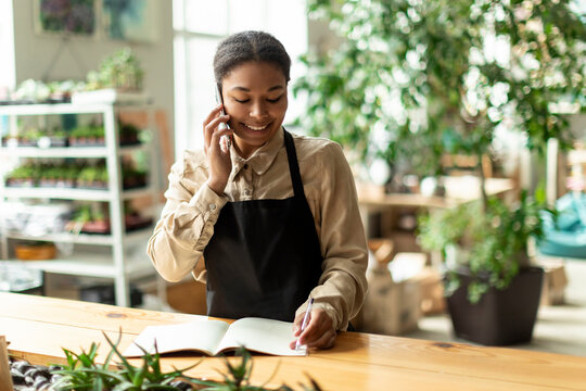 Black female gardener noting client order during mobile phone conversation, workin in greenhouse. Successful botanist and store owner concept