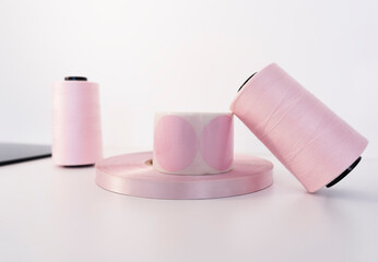 Pink coil of satin ribbon for labels or branding, roll of labels for thermal printing and two spools of thread. Clothing production.