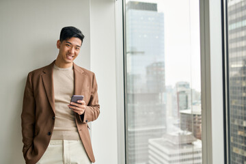 Young happy Asian business man executive using mobile cell phone tech standing in office at window....