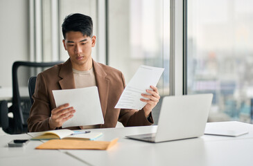 Young busy Asian business man lawyer, tax accountant manager holding paper documents checking bills, doing sales invoice accounting, reading legal contract or bank statement sitting at desk in office. - Powered by Adobe