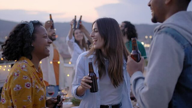Three happy multiracial young people dancing at rooftop party with friends in background. Excited persons toasting with beer at barbecue in evening. Colleagues celebrating weekend break outdoors.