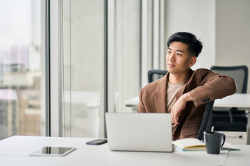 Thoughtful happy young Asian business man employee sitting with laptop in company office. Male professional manager executive looking at window thinking of future goals, dreaming or leadership.