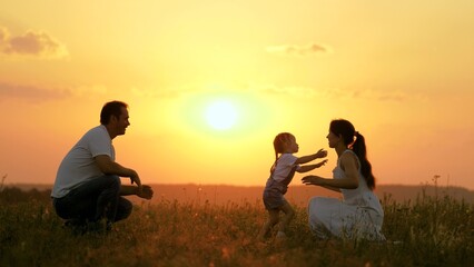Happy family walks in park at sunset. Mom, dad and baby. Little daughter goes from mom to dad, hugs and kisses her parents in rays of a warm sun. Healthy family plays in field. Happy family concept