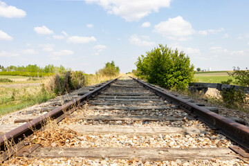 Fototapeta na wymiar traintracks of railway in the countryside with grass on either side and small bushes, blue cloudy sky