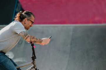 A happy urban tattooed middle-aged man is leaning on his bmx bike in a skate park and typing...