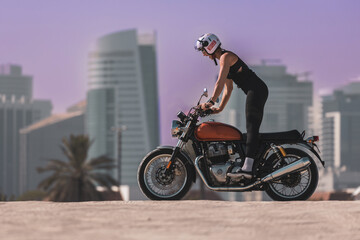 Fototapeta na wymiar woman driving a classic motorbike standing on foot stands and excited on a dubai beach with downtown in the background at sunset