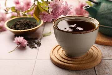 Traditional ceremony. Sakura petals in cup of brewed tea, teapot and flowers on tiled table, closeup