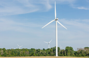 One large wind turbine and other turbines in a wind farm in the distance behind trees.