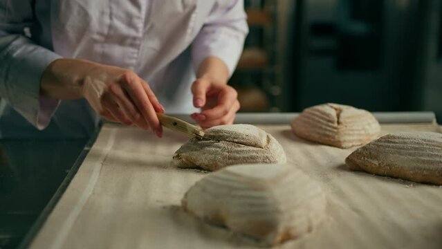 woman baker cuts patterns on raw bread rolls with a professional baker's knife before baking bakery production of pastries close-up