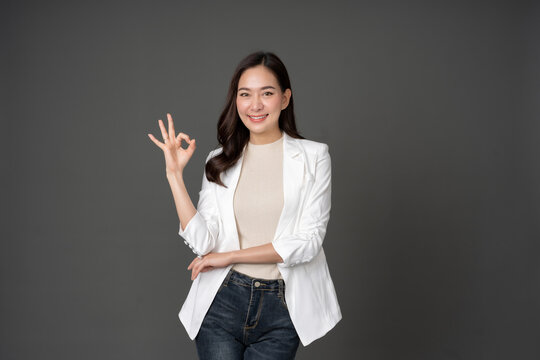Asian female executive with long hair Raise your hand and sign OK. Have a beautiful smile. wearing a white suit and stand to take pictures with a gray scene in the studio