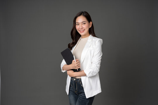 Asian female executive with long hair standing hugging a tablet for work, holding a pen, with a beautiful smile. wearing a white suit and stand to take pictures with a gray scene in the studio