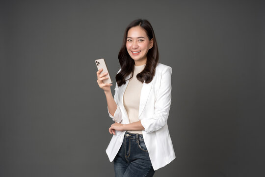 Asian female executive with long hair smile brightly and hold the phone confidently wearing a white suit Standing and taking pictures against the gray background in the studio