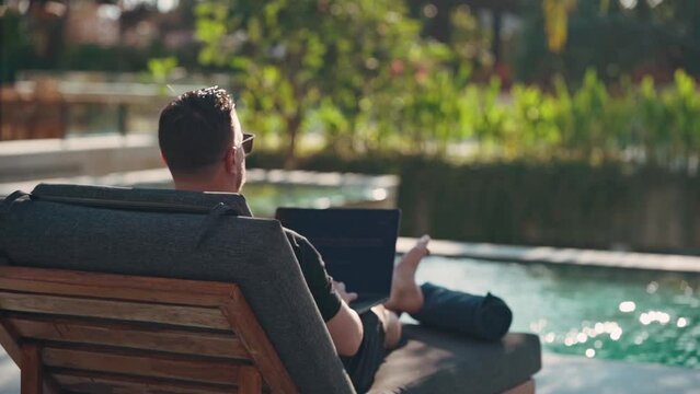 A man works at a computer in a beautiful villa on a sunny day. Remote work in front of the pool. The pleasure of working in a heavenly place.