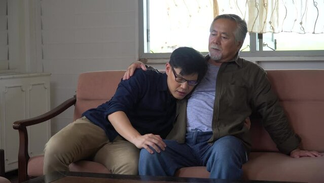 asian senior father embracing comforting pat back talking and laughing wirh adult  son on sofa in living room at home. happy family love .