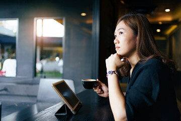 Happy cheerful Asian beautiful woman sitting in the coffee shop in front of the window and working on digital tablet and laptop - notebook computer.