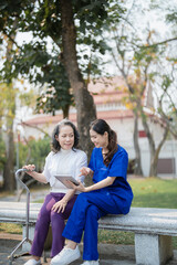 Patient care,, young Asian female nurse caring for with a cane and doing leg and knee exercises using a digital tablet as a teaching tool during a walk in the park parks.