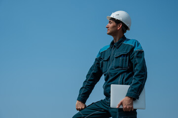 A male builder in a helmet stands against a blue sky with a laptop in his hands. 