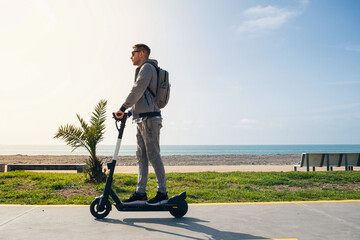 Young man traveler riding e-scooter along the sea coast and beach at sunny summer day. Male driving electric scooter outdoors. Sustainable lifestyle concept