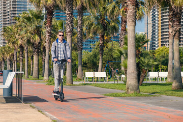 Young man riding electric scooter eco friendly transport along the promenade in the city with palm trees. Male driving e-scooter sustainable and outdoor in summer day. Sustainable lifestyle 