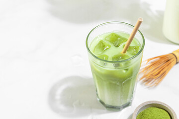 Glass of iced green matcha latte with a straw and bottle of milk on white tile background with hard shadows
