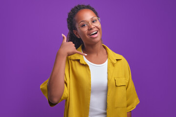 Young positive attractive African American millennial woman making call me gesture and smiling looking at camera inviting to talk on mobile phone after work stands on plain purple background.