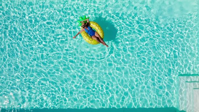 4K drone footage of beautiful woman dressed blue swimsuit floating on yellow inflatable pineapple ring in swimming pool. Resting, summer holidays, free time concept.